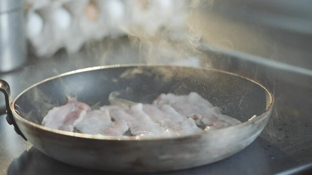 A pan of bacon is cooking on a stove, with steam rising from it. The bacon is sizzling and he is almost done