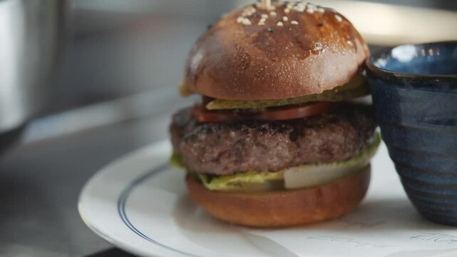A burger with a bun and a pickle spear on top of it. The burger is served on a white plate