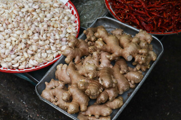 Market display with garlic cloves in a red-rimmed bowl, fresh ginger roots in a metal tray, and dried red chilies in another bowl, arranged at a traditional market stall.