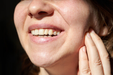 Obraz premium Close-up of young caucasian female smiling and touching cheek with healthy teeth in bright light.