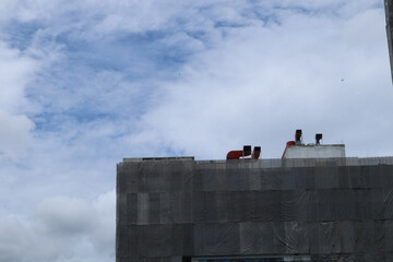 A building under construction or renovation is partially covered with protective mesh, with ventilation pipes and red exhaust ducts visible on the rooftop, set against a mostly cloudy sky.