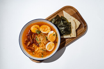 A comforting bowl filled with steaming noodles, perfectly boiled eggs, and rich toppings sits elegantly on a wooden tray. Accompanied by toasted bread and crispy seaweed, this meal warms the soul