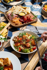 Plates filled with colorful delicacies surround a lively table. A hand delicately holds a fork, ready to savor a delicious dish while laughter and clinking glasses fill the air