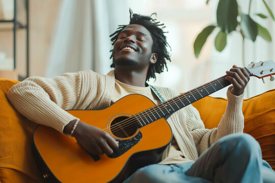 Young joyful african american man playing acoustic guitar at home, sitting on sofa in living room