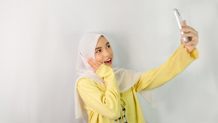 Beautiful and excited young Asian Muslim woman wearing hijab and yellow cardigan using smartphone to make video call or take selfie isolated on white studio background