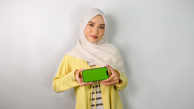 Young Asian Muslim woman wearing hijab smiling to camera while showing mobile phone with green screen while holding smartphone, promo video, gadget advertisement, online phone device.