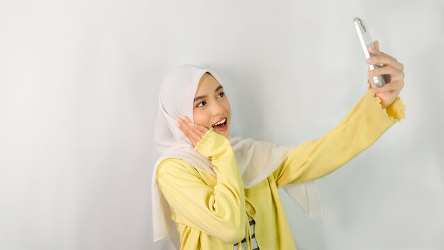 Beautiful and excited young Asian Muslim woman wearing hijab and yellow cardigan using smartphone to make video call or take selfie isolated on white studio background