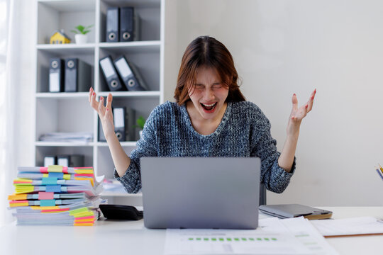 Portrait of young asia business woman sitting on desk with laptop under stress from too much work in the office.deadline job shouting crazy work overwhelmed, Mental Health in the workplace.
