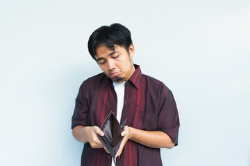 Asian Indonesian man showing his empty wallet with sad and worried expression. Isolated in gray...
