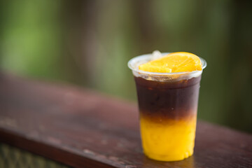 Plastic cup of iced coffee with orange juice and an orange slice on a wooden railing against a blurred natural background