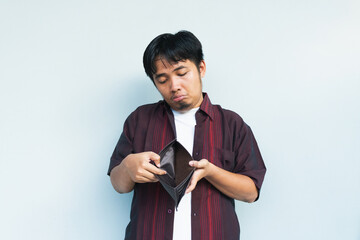 Asian Indonesian man showing his empty wallet with sad and worried expression. Isolated in gray...