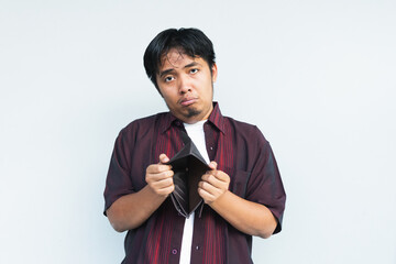 Asian Indonesian man showing his empty wallet with sad and worried expression. Isolated in gray...