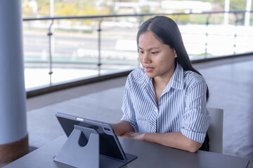 Young Asian woman working on tablet with stylus, focused and productive in a modern indoor/outdoor...