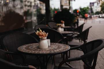 A charming cafe terrace with tables, chairs, and a vase of dried flowers adds a touch of charm awaits customers. An inviting outdoor cafe scene, with decorative tables and chairs, ready for patrons.