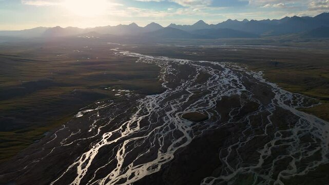 An expansive aerial view showcases a wide, braided river meandering through a vast valley with distant mountains under a setting sun