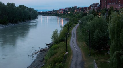 Serene riverside pathway during dawn or dusk with calm reflective water, warm lampposts, dense green foliage, urban cityscape, and distant mountains.