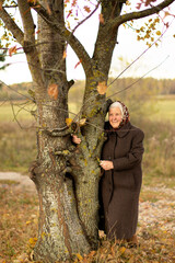 Old woman in coat and headscarf standing against the background of autumn trees