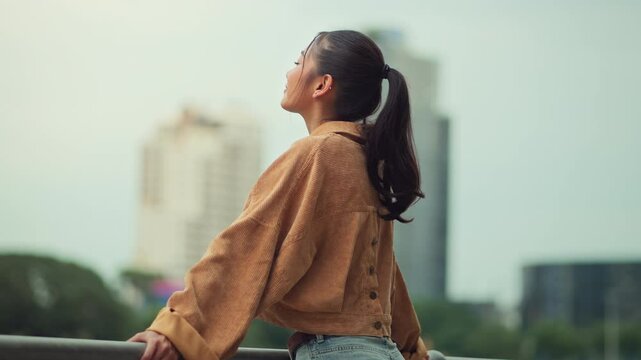 Cheerful asian woman standing on bridge in city park enjoying fresh air and nature &ndash; happy female smiling and breathing deeply in urban green space, mindful wellness lifestyle and outdoor relaxation