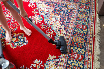 On a carpet with a colorful pattern, predominantly red and blue, human feet are visible as someone...