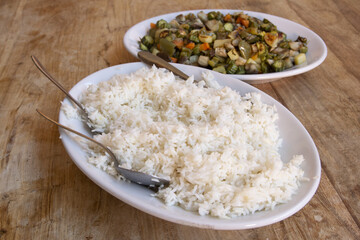 serving spoons resting on fluffy white rice next to cooked vegetables