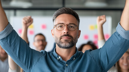 Obraz premium A confident man celebrates success with raised arms, colleagues smiling in the background at a collaborative workplace.