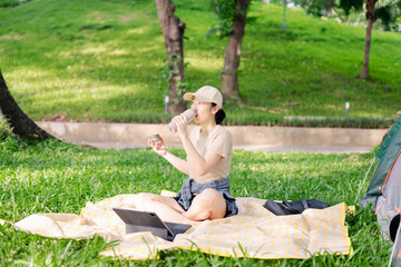 Young Asian woman enjoys a beverage while working on a laptop in a sunny park on a warm day