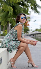 Stunning Asian woman sits confidently on a ledge by a serene canal, smiling, wearing a chic...