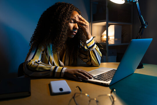 African American woman working late at night using laptop at home office having headache.