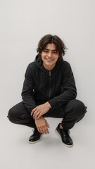 Full length of confident Spanish teenage boy in a zipped hoodie and track pants crouching slightly while smiling on plain white background
