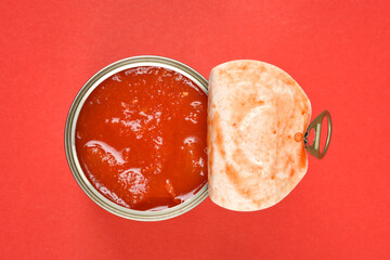 A top-down view shows an open can of chunky red tomato sauce with its lid peeled back, set against a vibrant red background
