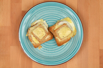 Two baked egg-in-toast with melted cheese and cooked eggs, served on a blue plate on a wooden...