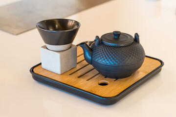 A traditional cast iron teapot and a black cup on a modern bamboo tray placed on a white surface