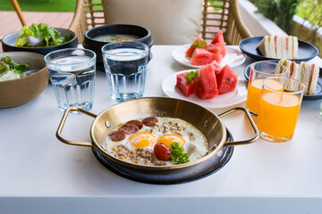 A delicious breakfast spread with fried eggs, sausage, salad, fresh fruit, sandwiches, and drinks on a white table.