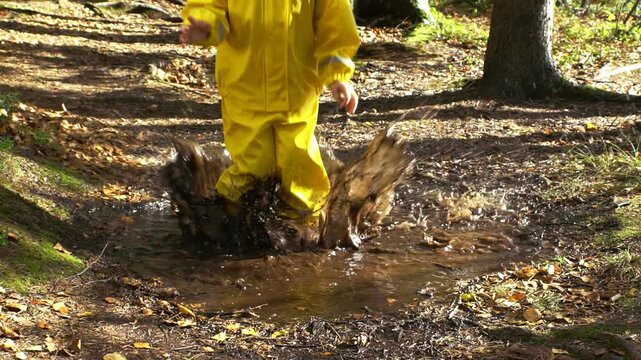 Happy child in yellow rain suit jumping in puddle in park on a sunny day. Active outdoor play, fun and nature exploration in Scandinavian woodland environment.