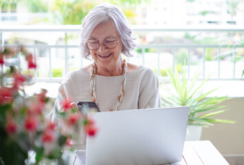 Smiling gray haired senior woman working on laptop sitting outdoor on terrace using smartphone. ...