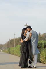 An affectionate Indian couple dressed in elegant traditional clothing shares a tender moment outdoors on a sunlit pathway