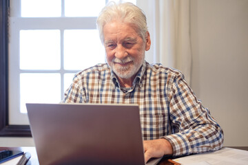 Fototapeta premium Confident mature man sitting at table with laptop and books working or studying. Attractive senior man enjoying teaching activity and learning using new technology