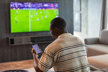 African man holding smartphone with sports bets app while watching football match at home in living room