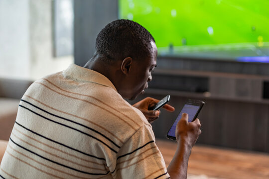African man holding smartphone with sports bets app while watching football match at home in living room
