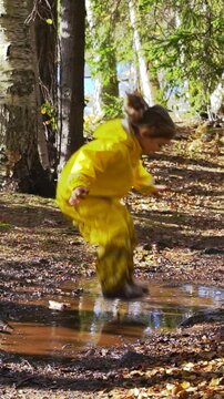 Happy child in yellow rain suit jumping in puddle in park on a sunny day. Active outdoor play, fun and nature exploration in Scandinavian woodland environment.