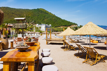 Sandy beach with wooden loungers, straw parasols and bar furniture in Icmeler, Turkey. Icmeler is popular coastal resort surrounded by green hills and calm sea under clear sky. 