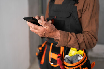 Close-up of builder hands with smartphone and belt with work tools in house interior. Concept of...