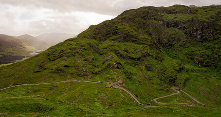 Vehicles traverse a winding road cutting into a lush green hillside in the Head of Gap of Dunloe. A lake and mountains are visible in the distance under a cloudy sky.