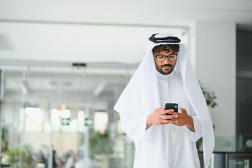 Arab businessman wearing traditional kandura using smartphone in modern office