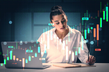 Businesswoman analyzing forex market data at night with digital chart overlays on laptop and notebook in fintech office environment.