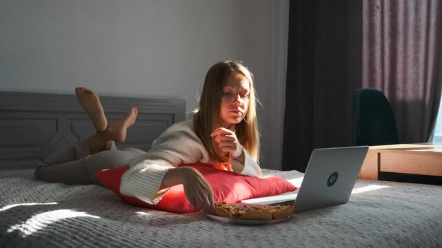 Casual work-from-home moment: woman in sweater using laptop while lying on bed savoring slice of pizza. Relaxed and efficient remote work setup.