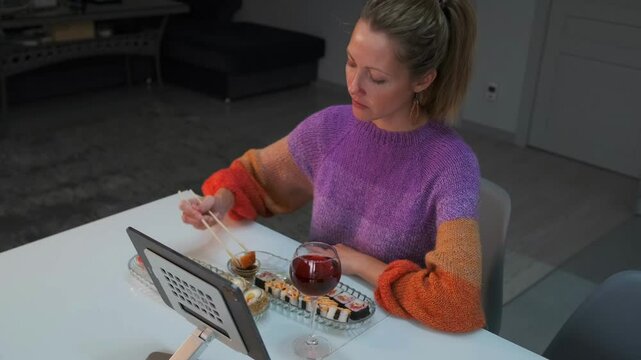 Young girl enjoying dinner delivered from Japanese restaurant eating sushi rolls dipping in soy sauce sitting in bright sweater in front of tablet next to glass of wine in modern living room.