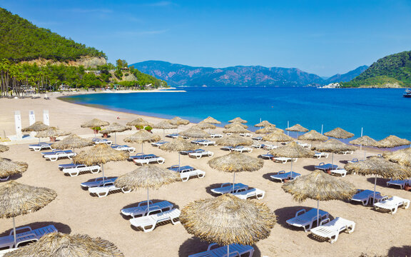 Sandy beach in Icmeler, Turkey, with rows of white sunbeds and straw parasols along turquoise shoreline. Icmeler is popular coastal resort surrounded by green hills and calm sea under clear sky. 