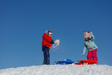 Two children in bright coats throw snow at each other on hilltop