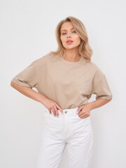 Blonde woman with wavy hair, wearing a beige short-sleeve top and white pants, poses confidently...